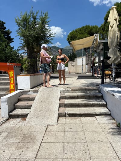 Two individuals are standing on a concrete ramp and steps in Budva, Montenegro, on a sunny day. The man, on the left, wears patterned swim trunks and a white towel wrapped around his head and shoulders. He is holding a small, white object in his hands and appears to be talking to the woman. The woman, on the right, is wearing a black bikini top and white shorts. She is holding a colorful beach ball and looking at the man. Behind them, to the left, is an orange structure with yellow letters spelling "BOX." To the right, there is a cafe with outdoor seating and umbrellas. In the background, there are trees and buildings, with a mountain range visible in the distance. The scene suggests a casual, outdoor setting likely near a beach or tourist area.