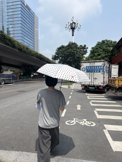A person walks away from the camera on a city street, holding a white polka-dotted umbrella over their head. They are wearing a grey t-shirt and black and grey striped pants. The person is walking on the sidewalk next to a road with traffic. In the background, there is a large modern building, a street lamp with multiple bulbs, trees, and a white truck with blue lettering. A crosswalk and a bicycle lane marking are visible on the road. The image appears to have been taken in Tianhe District, China, under a bright, cloudy sky.