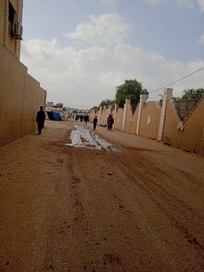 The image depicts a dirt road in Khan Yunis, Palestinian Territory, under a partly cloudy sky. The road surface is unpaved, showing tire tracks and a prominent muddy patch near the center of the frame. On the left, a light tan building wall with a textured finish extends into the distance, featuring an external air conditioning unit mounted high on its initial section. On the right, a decorative light tan wall with repetitive white or light-colored pillars and diamond-shaped inserts runs parallel to the road, topped by sections of metal fencing and partially obscured by green foliage. Numerous individuals, estimated at 8-10, are observed walking away from the camera along the road. One person in dark clothing is on the far left, near the building wall. Further down the road, a group of people, some appearing to carry bundles, are visible walking towards structures constructed from blue tarpaulins and other materials, suggesting temporary shelters or a camp in the middle distance.