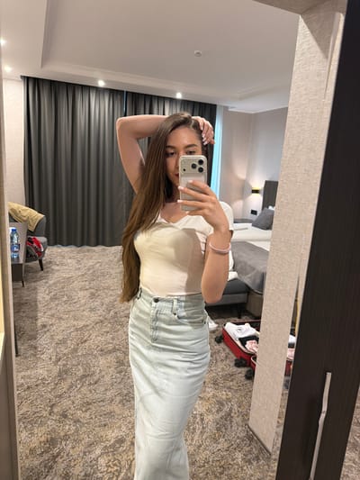 A woman is taking a mirror selfie in a hotel room. She is wearing a white t-shirt and light-wash jeans. She is holding her phone up to take the picture, with her other hand on her head. Her hair is long and brown. The room is dimly lit, with a large mirror on one wall. In the background, a bed, a dresser, and a window with dark curtains are visible. A suitcase and some other items are on the floor. The overall impression is that the woman is preparing to go out or has just arrived at her destination. There is no visible text. The photo appears to have been taken during the day, as natural light can be seen coming from the window, though the room itself is dimly lit. The setting appears to be a hotel room, likely in Hisarya, Bulgaria, given the provided context.