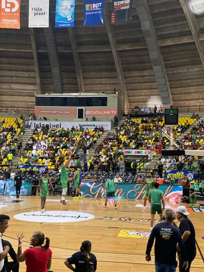 The image depicts an indoor basketball game in progress, likely within the Ciudad Deportiva Gral. Manuel Medina El Mejor in La Asuncion, Venezuela, given the presence of "Venezuela" on the court. The scene is viewed from a lower perspective, capturing a section of the stands filled with spectators in yellow seats, many wearing green. On the court, several male basketball players are engaged in the game, predominantly dressed in green uniforms. One player in the foreground is in the process of shooting a basketball. The arena features large banners hanging from the ceiling, advertising various companies and services, including "DATALINK," "Isla pixel," "CARIBEST WATER," "BDT," "ELEVATION 58 FITNESS," and "APUESTASROYAL.COM." Another banner reading "BNC BNC BNC" is visible in the stands. The lighting appears to be artificial, typical of an indoor sports venue. Several people are also visible in the foreground, some watching the game and others potentially involved with the event.