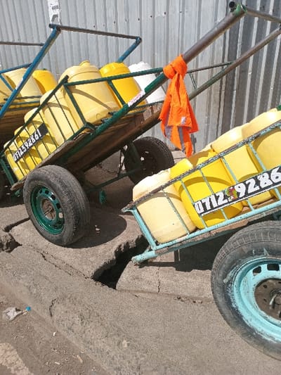 The image displays two two-wheeled handcarts, each loaded with multiple yellow plastic jerrycans, positioned on a damaged concrete surface adjacent to a corrugated metal wall in Nairobi, Kenya. The left cart features a dark green metal frame and green wheel hubs, carrying several yellow jerrycans. A black identification plate on this cart displays partially visible white numbers, including '0712 92861'. The right cart, partially visible, has a light blue metal frame and light blue wheel hubs, also laden with yellow jerrycans. An identification plate on this cart clearly shows the numbers '0712 9286'. An orange reflective safety vest is tied to the handle of the right cart. The concrete ground exhibits significant cracks and a pronounced gap between the two carts, with a curb present in the foreground. The background consists of a vertical corrugated metal sheet wall or fence. No individuals are present in the scene.