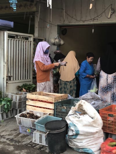 The image depicts an outdoor market scene in Sentul, Indonesia, likely a local produce stall. In the foreground, several crates and baskets are filled with various produce, including what appears to be taro roots in a white plastic basket, tomatoes in a wooden crate, and leafy green vegetables in grey plastic crates stacked behind. A large white sack, possibly containing goods for sale or delivery, dominates the right side of the frame.
Several individuals are visible at the stall. A woman in a light pink hijab and a burnt orange cardigan is standing to the left, holding a plastic bag with a product inside. To her right, another woman in a beige hijab is partially visible. Further back, a woman wearing a blue long-sleeved dress and a black head covering is interacting with the stall, holding a plastic bag. A fourth individual, wearing a dark hijab, is on the far right. The background shows the entrance to a building or stall, with various items hanging and stored within. The ground is paved and appears wet, suggesting recent rain or cleaning. The overall atmosphere is that of a busy marketplace.