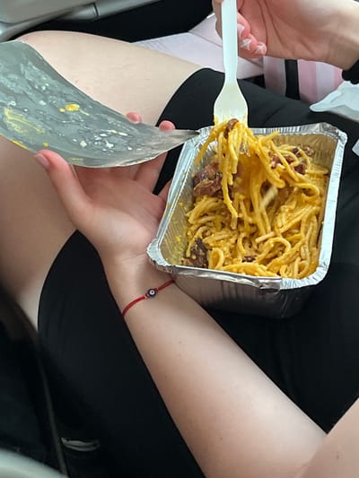 A person is shown seated, holding a rectangular aluminum container filled with spaghetti carbonara. The spaghetti is yellow and has pieces of meat mixed in. They are using a white plastic fork to eat the pasta. The person also holds the lid. The individual has pale skin, and is wearing a black outfit. A red string bracelet with a blue and black bead is visible on their wrist. The location is most likely Verona, Italy, based on the typical food choice. The scene suggests an indoor setting, possibly a restaurant or home.