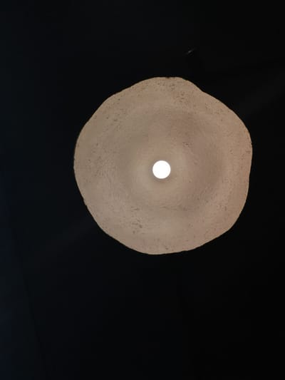 The image shows a close-up, low-angle view of a textured, off-white lampshade hanging from a dark ceiling. The lampshade is irregularly shaped, resembling a rough, organic form. A bright, circular light source is visible in the center of the lampshade, casting a warm glow. The background is predominantly black, with the exception of the lampshade and a faint, light-colored streak in the upper right portion of the image, possibly a reflection or a distant object. There are no people or animals present. The scene appears to be indoors, in an environment with dim ambient lighting. No specific location in the city of Кварса, Russia, can be determined from the image.