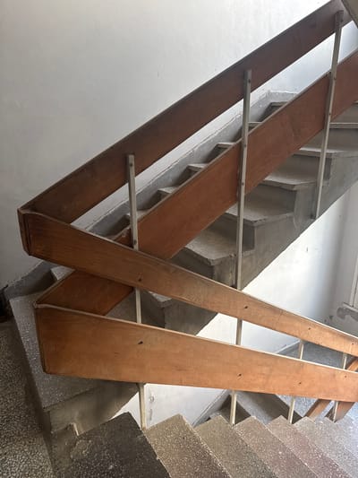 The image displays an interior view of a concrete staircase, located in the city of Sofia, Bulgaria. The perspective is from an elevated position, looking downwards across multiple flights of steps and an intermediate landing. The stair treads and risers are composed of light grey concrete with an exposed aggregate finish, showing evidence of wear and light surface debris. The structural underside of the staircase is raw, darker grey concrete. A distinctive railing system is present, consisting of two parallel, horizontal wooden planks, stained a reddish-brown color, which run along the incline of the stairs. These wooden planks are supported by slender, vertical, light grey metallic posts, secured with visible fasteners. The adjacent walls are painted a plain off-white or light grey, exhibiting minor scuffs, dirt, and textural inconsistencies. The visible lower landing features a matching aggregate concrete surface. No individuals, actions, or dynamic interactions are depicted in this static architectural scene.