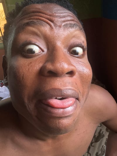 The image is a close-up selfie of a Black individual with their tongue sticking out. The person's eyes are wide open, and their forehead is wrinkled. The skin appears to have some texture and a slight sheen, possibly due to sweat. The background is dark and out of focus, with hints of a brown surface on the right and a lighter, possibly patterned fabric on the lower right. The image is taken in Ikorodu, Nigeria.