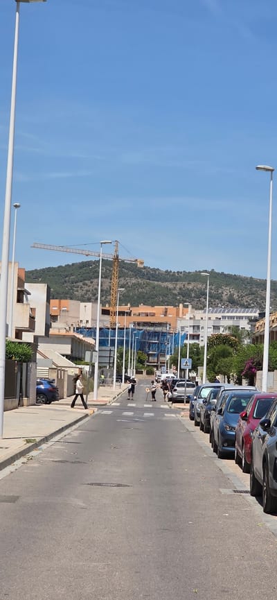 The image depicts a street in Sagunto, Spain, lined with parked cars on the right side. A construction crane is visible in the background, behind a row of buildings. Several people are walking in the street, and one person is walking on the sidewalk. Streetlights line the left side of the road, extending towards the sky. Hills covered with vegetation are in the distance. The sky is clear and blue.