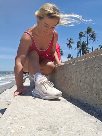 A young blonde woman in a red swimsuit is crouching on a concrete embankment in Mui Ne, Vietnam. She is looking down at her right foot, which is clad in a beige athletic shoe and white socks. Her hair is blowing in the wind. In the background, the ocean and palm trees are visible under a clear blue sky. The embankment appears to be part of a coastal walkway or sea defense. The lighting suggests it is daytime, likely mid-morning or afternoon given the bright sunlight and shadows.