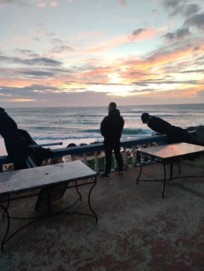 The image displays an outdoor terrace scene at twilight, overlooking the sea in Tanger, Morocco. A single male subject stands with his back to the camera, observing the ocean. He is dressed in a dark, possibly hooded, jacket, dark trousers, and athletic shoes. He stands behind a blue railing with white balusters separating the terrace from the sea. The ocean exhibits moderate waves breaking against a visible rocky shoreline. The sky above is characterized by twilight colors, featuring hues of pink, orange, purple, and blue, with scattered clouds. The foreground terrace is made of concrete and shows patches of discoloration. Two square tables with light-colored tops and dark, ornate metal bases are positioned on the terrace. A dark, covered object, possibly a folded chair, is present near the table on the left. A second similar dark object is partially visible to the right of the standing subject, positioned behind the rightmost table.