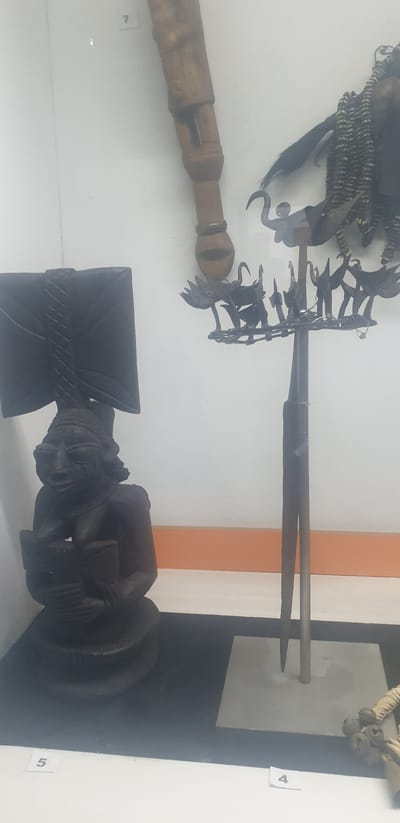 The image shows an interior display, possibly in a museum, presenting cultural artifacts. In the foreground is a dark wooden carving of a figure with a stylized axe. Above it is a collection of objects, including a spear-like item with a metal shaft and a decorative, possibly ceremonial, headdress or adornment. A wooden handle is visible beside the headdress, possibly related to the artifacts. The number tags "4", "5", and "7" are visible next to the artifacts, which likely identify them. The location is Vom, Nigeria. The items are positioned against a white and orange wall.