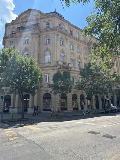 The image depicts a street corner in Budapest, Hungary, with a prominent building on the left and a tree-lined street on the right. The building is a multi-story structure with classical architectural elements, featuring ornate decorations, arched windows, and balconies. The lower facade of the building includes storefronts. On the street in front of the building, there are pedestrian crossings and yellow road markings. Two individuals are walking on the sidewalk, and a red fire hydrant is visible. Further down the street, a black car is parked, and a few trees are present. The sky is blue with scattered clouds. The overall scene suggests an urban environment on a sunny day.