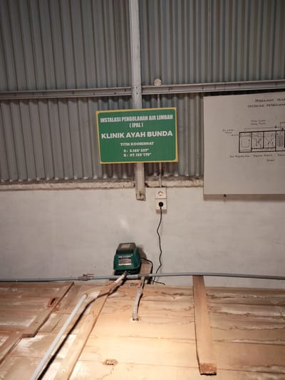 The image depicts the interior of a facility in Lhokseumawe, Indonesia, likely an industrial or processing plant, characterized by corrugated metal walls and a concrete ceiling. Dominating the center is a green sign indicating "INSTALASI PENGOLAHAN AIR LIMBAH (IPAL) KLINIK AYAH BUNDA" with coordinates S: 5.185° 537' E: 97.135° 175". To the right, a diagram titled "PENGAJUAN DESA INSTALASI PENGOLAHAN AIR LIMBAH" illustrates a wastewater treatment system, labeling components such as "Air Limbah Masuk," "Media Plastik Sarang Tauen," "Bak Pengendap Awal," "Biopilter Anaerob," and "Aerator."
On the wall below the sign, a white electrical outlet with two sockets is visible, with a black power cord plugged into the lower socket and extending downwards. In the lower portion of the image, a green air pump or blower is placed on a wooden platform. Gray pipes, some with valves, are routed above and across this platform, connecting to the blower and disappearing out of frame. The wooden platform appears to be a part of a structure that is stained and weathered, suggesting it may be part of a tank or processing unit. The overall lighting suggests an indoor environment with artificial light sources. There are no people visible in the image.