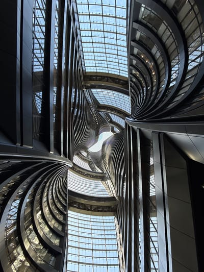 The image is a low-angle view of the interior of a modern building. The architecture is characterized by sweeping curves and lines, creating a vortex-like effect towards a central skylight. The building's structure is predominantly constructed of metal and glass, with horizontal lines suggesting multiple levels or floors. The glass allows natural light to permeate the interior. The scene is located in the Fengtai District of China.