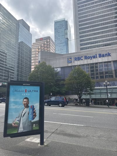 The image shows a street scene in downtown Vancouver, Canada. A billboard in the foreground displays an advertisement for Michelob ULTRA Zero, featuring soccer player Lionel Messi holding a can and a soccer ball. Behind the billboard, several modern high-rise buildings dominate the skyline, including one with "RBC Royal Bank" signage. The street is paved with asphalt and has visible lane markings and a yellow center line. A dark blue SUV is parked on the side of the road, and several pedestrians are walking on the sidewalk in front of the RBC Royal Bank building, where a McDonald's restaurant is also visible. The sky is overcast, suggesting a cloudy day. The overall environment appears to be an urban business district with a mix of commercial buildings and public infrastructure.