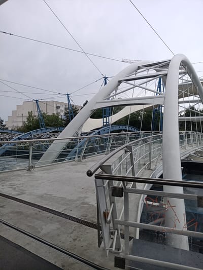 The image shows an exterior view of the new pedestrian bridge connecting the Holešovice and Karlín districts over the Vltava river in Prague, Czechia. The bridge features a distinctive white arched structure and is supported by a network of cables and blue steel elements. The walkway is made of concrete with metal railings and glass panels. The sky is overcast. There are no people visible in the image, but there is some red graffiti on a glass panel of the railing in the foreground. In the background, a building and trees are visible.