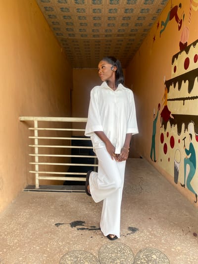 A young Black woman stands in a narrow, covered walkway with orange-painted walls. The ceiling is decorated with a repeating blue and white floral pattern. On the right wall, a colorful mural depicts a multi-tiered cake with animated figures. A white railing with horizontal bars is visible on the left. The floor is made of speckled terrazzo. The woman wears a white, loose-fitting shirt and pants. She is looking to her left, with her left leg slightly bent. Her hands are clasped in front of her. There are dark stains on the floor. The overall impression is that of an outdoor public space, possibly near a school or community center in Gura Top, Nigeria.
