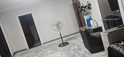 The image depicts an interior scene, likely a living room, located in Uychi, Uzbekistan. A tall, oscillating fan stands in the center of the room, its base on a patterned carpet. To the left, a dark wooden door is slightly ajar, revealing a glimpse of another room. On the right side of the room, a plush armchair is visible, partially obscuring a small table. On this table sits a blue vase with dark red artificial flowers, a white air freshener, and another white electronic device. Further to the right, a desk with a computer monitor, keyboard, and mouse is partially visible. The walls are covered in a textured, light-colored wallpaper, and the ceiling has white molding. The lighting suggests it is daytime, with natural light possibly entering from an unseen window. There are no people present in the image.