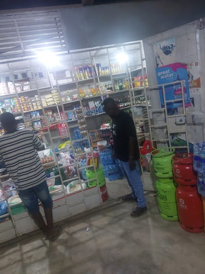 The image shows the interior of a small store, likely in Dar es Salaam, Tanzania, given the brands of gas cylinders and water bottles visible. Two men are visible in the store. One is wearing a black t-shirt and jeans and appears to be examining items on the shelves. The other man is wearing a striped shirt and jeans, also looking at items on the shelves. The store is well-stocked with a variety of goods, including drinks, food items, and gas cylinders. The shelves are made of metal and are organized in a grid pattern. The lighting in the store is bright. Gas cylinders, some red with the "ORYX" logo and some green with the "TAIFA GAS" logo, are stacked on the top shelves. Bottled water with the "Great Water" brand is also prominently displayed.