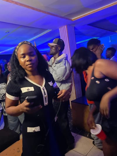 This image depicts a dimly lit indoor scene, likely a party or gathering, with vibrant blue lighting illuminating the space. The setting appears to be a venue in Durumi, Nigeria.
In the foreground, a woman is the central subject, holding a black smartphone in her left hand. She is wearing a black short-sleeved dress adorned with white trim and decorative buttons. Her hair is dark and curly, and she is looking towards the camera with a neutral expression.
Behind her, to the left, a man wearing a light-colored hooded jacket and a baseball cap is visible. Further back, another individual's face is partially obscured by other people. To the right, a woman in a black tank top with decorative elements is captured in motion, her body turned away from the camera. A red plastic cup is held in her hand, suggesting a social event. The background features striped columns and patterned walls, typical of entertainment venues. The floor has a tiled pattern. The overall atmosphere is one of a lively social gathering.