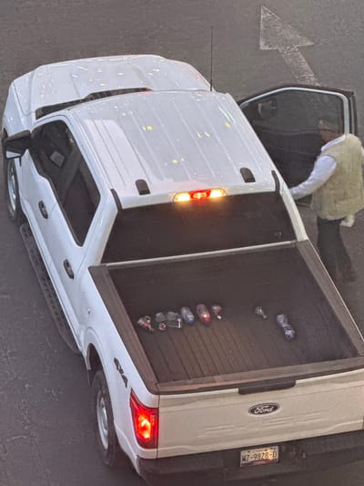 A top-down view shows a white Ford pickup truck parked on asphalt. The truck's tailgate is down, revealing several empty water bottles and a small bottle inside the bed. A person is partially visible near the open driver's side door. An arrow marking a traffic lane is painted on the asphalt near the front of the truck. The license plate reads "WZ-9828-D" and indicates Tlaxcala. "4x4" is printed on the rear of the truck. The scene is located in San Bernardino Tlaxcalancingo, Mexico.