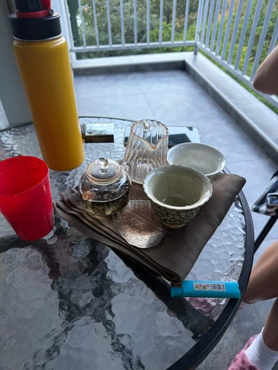 The image shows a table setting on a balcony or patio. The table is made of glass with a textured surface. On the table are several items related to tea drinking, including two white teacups, one with a blue floral pattern and cracks, a clear glass teapot filled with tea leaves, a clear glass pitcher, and a small decorative metal leaf. A brown cloth napkin is folded beneath the teacups and teapot. A bright blue disposable lighter with warning symbols and a barcode is also on the table.

In the foreground, a large yellow water bottle or thermos and a red cup are visible. In the background, parts of a person's legs, wearing white socks and fluffy pink slippers, are seen, suggesting someone is seated nearby. The scene appears to be outdoors, with a slatted railing and greenery visible through it, indicating a balcony overlooking a natural environment. The lighting suggests it is daytime. There is no specific indication of the weather or time of day beyond daylight.

The overall impression is a casual outdoor setting, possibly for a tea break. The items are arranged as if in use or about to be used. The presence of the lighter and the water bottle suggests a relaxed, possibly informal gathering. The location context of Tbilisi, Georgia, is provided but not visually discernible from the image itself.