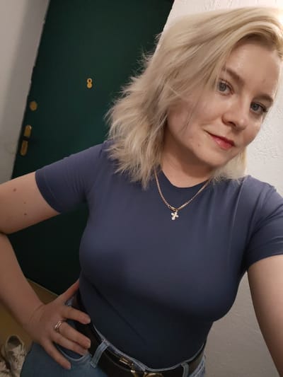 A blonde woman with blue eyes and light makeup is taking a selfie. She is wearing a dark blue t-shirt, blue jeans, a belt, and a gold necklace with a cross pendant. She has a ring on her left hand. The background appears to be an indoor setting with a dark green door and a light-colored wall. There is no indication of the location being in Izhevsk, Russia.