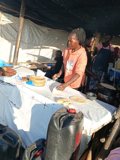 Under a makeshift shelter, a woman in a peach hoodie rolls dough on a white-clothed table, surrounded by prepared chapattis and sweet potatoes. Two other women, one in an apron, are visible behind her. In the background, a man sits with a cup. Fuel canisters are stacked in the foreground, and a cooking pot sits on a burner to the right. The scene conveys a busy outdoor kitchen or street food operation.