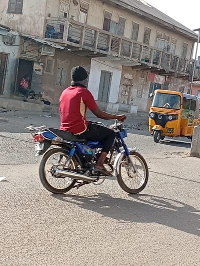 The image captures an urban street scene in Gawuna, Nigeria, under bright daylight. A dark-skinned individual, appearing male, is centrally positioned on a blue and silver motorcycle, facing away from the viewer. The person wears a red short-sleeved shirt with light-colored side panels, dark trousers, and a black head covering that also functions as a face mask. Bare feet, clad in flip-flops, are visible on the motorcycle's footpegs. The motorcycle features spoked wheels, silver front and rear fenders, a chrome exhaust pipe on the right side, and a silver luggage rack over the rear wheel.
In the background to the left, a two-story building with a weathered, light-colored facade is visible. Its ground floor features multiple doorways, one of which is open, revealing a second individual, appearing male, seated on a wooden bench inside. This person is wearing a red short-sleeved shirt and dark trousers. Above the open doorway, a white sign with black text is mounted. The building's first floor includes a long, open balcony with a railing and multiple windows, some with closed shutters.
To the right of the motorcycle, a yellow motorized rickshaw, commonly known as a Keke Napep, is positioned on the road, facing partially away from the camera. The number "3958" is visible on its rear panel. A head is discernible within the rickshaw, suggesting a driver or passenger.
The ground surface is a textured, light-colored pavement, consistent with an urban road. Distinct shadows of the motorcycle and its rider are cast onto the pavement, indicating strong overhead sunlight.
