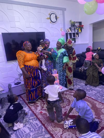 This image captures an indoor gathering in the city of Kwang, Nigeria. In the central area, four adult women in traditional Nigerian attire, including headwraps and patterned dresses, are clustered together. One woman, wearing a blue and purple patterned outfit, holds a young child dressed in a colorful patterned outfit. The child appears to be interacting with a pink, multi-tiered birthday cake, adorned with princess motifs, positioned on a low table directly in front of the group.

On the floor in the foreground, three young boys are present: one boy in a black t-shirt crouches on a grey tiled floor, another boy in a white t-shirt stands facing the cake, and a third boy in a light blue shirt kneels on a red patterned rug, engaging with a white and blue toy truck. In the background, on a black leather sofa, at least two more children are visible; one child in a pink shirt stands on the sofa, and another in a green dress sits.

The room features a white wall with a textured wave pattern, mounting a large flat-screen television and a gold-rimmed wall clock. Pink, green, and purple balloons are visible near the ceiling in the background. A dark, compartmentalized wall shelf is also present. The floor combines grey marbled tiles and a large red rug with intricate patterns. The scene is illuminated by interior lighting.