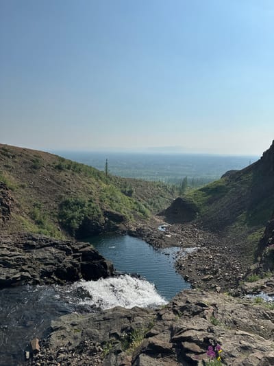 The image depicts a scenic natural landscape with a waterfall cascading into a pool of water. The foreground is dominated by dark, jagged rocks, with a foamy white waterfall spilling over the edge. The water then flows into a calm, dark blue pool. Steep, barren hillsides flank the pool, covered in loose rocks and sparse vegetation. Further in the distance, the landscape opens up to a vast expanse of forested land, shrouded in a hazy atmosphere, suggesting a considerable distance. The sky above is a clear, pale blue, transitioning to a lighter hue near the horizon. The scene appears to be a natural reserve or park, possibly located near a city, as evidenced by the distant urban or semi-urban sprawl visible through the haze. The presence of a few people swimming or wading in the pool suggests it is a popular recreational spot. The overall impression is one of rugged natural beauty and a sense of vastness. The location is identified as Talnakh, Russia.