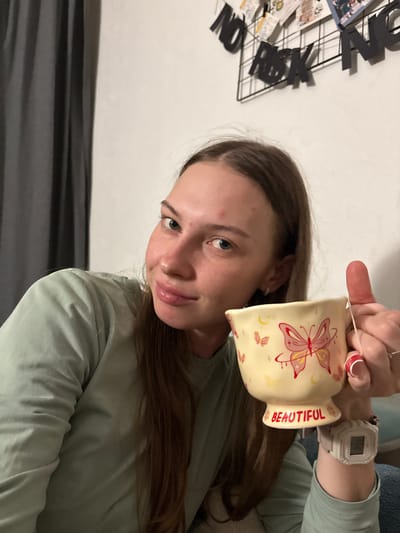 A young woman with long brown hair and a light green long-sleeved shirt is holding a yellow mug with a butterfly design. The mug has the word "BEAUTIFUL" written on it. She is wearing a white digital watch on her left wrist. The background is a white wall with a black wire grid on it, decorated with letters and photos. A dark grey curtain is visible on the left side of the image. The scene appears to be indoors, likely in a residential setting, and no specific location in the city of Perm, Russia, can be determined from the visual information.