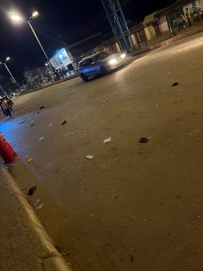 The image depicts a nighttime street scene in Bukuru, Nigeria. The street is paved and wet, with scattered debris, including papers and dark, possibly plastic, trash bags. Streetlights illuminate the scene, casting a warm glow on the wet pavement. In the background, buildings line the street, some with illuminated signs and windows. A blue car is visible in motion, its headlights on, suggesting it is either driving or has just passed by. To the left, a red cylindrical object, possibly a traffic cone or a painted pole, stands near the curb. Another object, possibly a motorcycle or a tricycle, is partially visible on the far left. The overall impression is of a busy urban street at night, likely after rain, with evidence of pedestrian and vehicular activity.