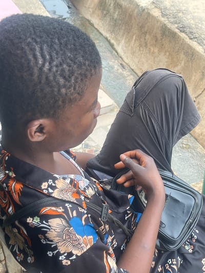 A person, likely a young woman, is sitting outdoors on a concrete bench. She has dark skin and short, dark hair styled in waves. She is wearing a black dress with a floral print featuring orange, blue, and beige flowers. A black crossbody bag with a zippered compartment rests on her lap. Her left hand is visible, resting on her leg. The background consists of weathered concrete structures and some water, suggesting an outdoor public space. The scene is captured in a medium shot, focusing on the individual and her immediate surroundings.