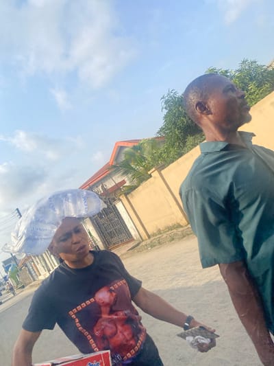 The image depicts an outdoor daytime street scene in Iranla, Nigeria. In the foreground, a dark-complexioned woman is captured from the chest up, positioned slightly left of center, wearing a black short-sleeved t-shirt with a graphic of a shirtless male figure. She carries a clear plastic bag containing multiple sachets of "PLOM" brand drinking water balanced on her head. In her left hand, she holds a red and white box, identifiable as "Indomie" noodles. Her expression is focused, and her gaze is directed slightly downwards and to her right. To the right and slightly behind the woman, a dark-complexioned man is seen in profile, looking upwards and to the right. He wears a dark teal short-sleeved collared shirt and exhibits visible perspiration on his face and arm. In his left hand, he holds a small, dark, folded object. The background reveals a light-colored, unpaved road, a tan-colored wall, green foliage including trees and bushes, and a building with an orange-red gabled roof and a dark metal gate. The sky is light blue with scattered white clouds. Power lines are partially visible on the far left. The two individuals appear to be in transit, but no direct interaction between them is evident.