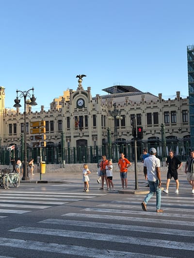 In front of the Estació del Nord, a historic train station in Valencia, Spain, people are crossing a street at a pedestrian crossing. The station, a prominent building with ornate architecture and a large clock, serves as the backdrop. Several individuals are captured in motion, some walking across the zebra stripes, while others are standing or conversing on the sidewalk. The sun is casting long shadows, indicating it is likely late afternoon or early morning. The scene is bustling with activity, typical of a public space in a city.
