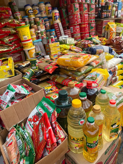 A variety of grocery items are densely packed. Canned goods, including tomato paste and tuna, are piled on top of mayonnaise jars. Packets of spaghetti, seasoning, and instant noodles are interspersed with cooking oil bottles. Boxes filled with packets of "Gino" tomato seasoning are visible. Brand names like "SONA," "LAZIZ," "KING'S," and "BENNY" are visible on various products. The arrangement suggests a bulk purchase or stock storage.