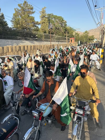 A large group of men on motorcycles are lined up on a paved road. Most of them are holding Pakistani flags. A mountain is visible in the background. A barrier with barbed wire is on the left. The location is likely Quetta City Tehsil, Pakistan.