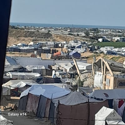 The image displays a sprawling, densely packed displacement camp in Khan Yunis, Palestinian Territory, under clear daylight conditions. Numerous temporary shelters and tents, constructed from various materials including white, light blue, grey, and brown tarpaulins and fabrics, dominate the foreground and midground. Some structures incorporate visible wooden frames. The camp is situated on sandy, undulating terrain, with areas of cleared ground and low concrete walls visible. In the midground, a prominent orange tarp-covered object is discernible among the shelters. Beyond the immediate settlement, the landscape transitions into sandy hills, which meet a coastal strip featuring scattered low-rise buildings. The Mediterranean Sea stretches across the distant horizon, beneath a clear blue sky. No specific individuals, actions, or interactions are clearly identifiable within the frame due to the distance and resolution.