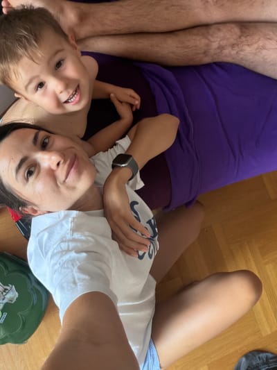 A high-angle, top-down shot captures a cozy scene of two people, likely a mother and her young son, resting together on a wooden floor. The son, positioned closest to the camera, beams with a wide, joyful smile, his eyes bright and his mouth showing teeth. He is wearing a white t-shirt with a blue and white pattern. He rests his head on his mother's chest, who is lying beside him. Her expression is relaxed and content as she looks towards the camera, one arm resting on her son's back, the other around him. She is wearing a white t-shirt. A smartwatch is visible on her left wrist. The son's leg is visible on the left side of the frame, with the mother's leg extending to the right, wearing what appears to be blue and white striped shorts.
Behind them, a large swatch of vibrant purple fabric, possibly a blanket or clothing, provides a colorful contrast to the wooden floor. A dark green, dinosaur-themed object, possibly a toy box or case, sits on the floor near the mother's legs. The overall mood is one of warmth, love, and familial connection. The natural lighting suggests it could be daytime. No specific time of day, weather, or explicit location cues beyond the wooden floor and the general context of Budva, Montenegro are discernible.