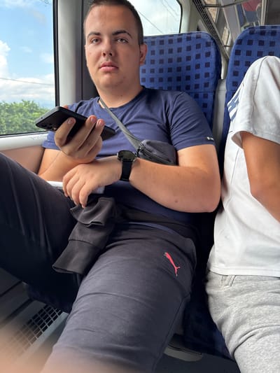 The media file shows two people sitting on a train. The person in the foreground, a young male, is holding a smartphone and looking towards the camera with a neutral expression. He is wearing a navy blue t-shirt and dark pants with a red Puma logo on the leg. A black bag is slung across his chest, and he has a watch on his left wrist.

Behind him, another person, whose face is not fully visible, is wearing a white t-shirt and grey sweatpants. Both individuals are seated on blue train seats with a checkered pattern. Through the window, a bright sky with clouds and green trees can be seen, suggesting it is daytime with clear weather.

The image is oriented sideways, and the overall impression is of a casual journey. The scene appears to be set on a public transport vehicle, likely a train, given the seating arrangement and the interior details. The context provided indicates the location is Plovdiv, Bulgaria. No visible text is present in the image.