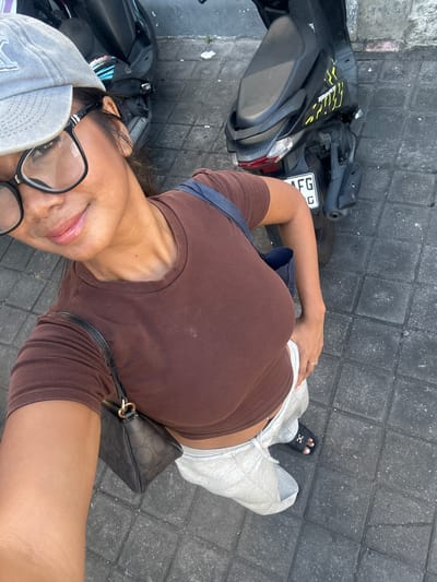 A person is taking a selfie while standing next to a parked scooter on a paved sidewalk. The person is wearing a brown crop top, grey sweatpants, black sandals, a grey baseball cap, and glasses. A black handbag with a repeating pattern and gold hardware is slung across their body. The scooter is black with yellow decals and a license plate that reads "FG C". The paved ground is made of dark grey interlocking bricks. The lighting suggests it is daytime, possibly late afternoon or early morning due to the soft shadows. The person appears relaxed and is smiling slightly at the camera. The scene is set outdoors on a street in Denpasar, Indonesia.