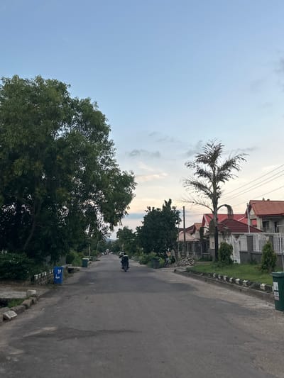 The image depicts a street scene in Abuja, Nigeria, likely in a residential area. A paved road runs through the center, bordered by green vegetation and houses with red roofs. On the left side of the road, a large, leafy green tree dominates the foreground. Further down the street, a motorcycle with a rider is visible, moving away from the camera. Several houses with white fences and neatly manicured lawns are present on the right side. A few people are casually seated near some rocks on the left side of the road, and two blue and green trash bins are placed along the roadside. The sky above is a soft blue with scattered clouds, suggesting it is either early morning or late afternoon. The overall impression is one of a quiet, suburban environment.