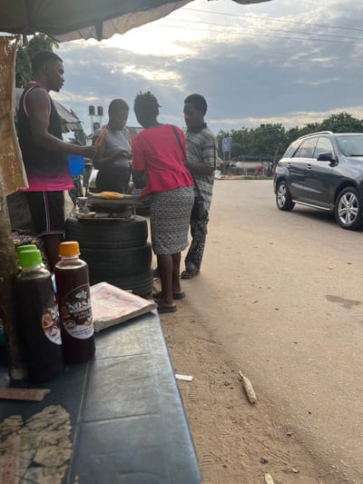 The image depicts an outdoor scene on a paved road, likely in Abuja, Nigeria, under a cloudy sky. In the foreground, a dark blue table is visible with two bottles of dark liquid, one with a green cap and another with an orange cap, and a closed book. The table is positioned to the left of the frame. To the right of the table, a group of four individuals is gathered.
Three individuals, appearing to be a man and two women, are standing around a makeshift grill. The grill consists of corn being roasted on a wire rack placed on top of a stack of tires. One woman, wearing a bright pink top and a black and white patterned skirt, is facing the grill. A man in a black and pink tank top and black shorts is to her left, holding a blue fan. Another woman, wearing a grey top, is to the left of the grill, looking down. A fourth individual, a man in a striped t-shirt and patterned trousers, stands behind the woman in the pink top.
Further to the right, a dark-colored SUV is parked on the side of the road, facing away from the camera. The background features trees and some low-lying structures, possibly shops or stalls. A single cob of corn is lying on the ground to the right of the road. The overall impression is of a street-side food vendor operation.