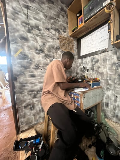 The image depicts a young Black man, likely in his late teens or early twenties, wearing a light brown t-shirt and dark pants, seated at a makeshift workbench. He appears to be engaged in repairing electronic devices. The workbench is cluttered with various electronic components, wires, tools, and a small blue electronic device. Behind him, on a shelf, are packaged electronics, including a "BASS" branded item and another box with "HOT" printed on it. A handwritten note is attached to the wall, listing an account number, a name "SAKA JAMILU," a phone number "09067076145," and "Engineer Opeyemi." Another smaller, handwritten note to the left advises customers to leave phones with the cashier and states the business is not responsible for loss or damage.

The environment appears to be a small, somewhat disorganized workshop or repair shop. The walls are covered in a gray and white mottled material, possibly painted or wallpapered. The floor is a mixture of dirt and possibly water, suggesting an outdoor or semi-outdoor setting, common in areas experiencing rain. In the background, another person in a similar t-shirt is visible, and a white plastic chair is present. Various other electronic items, including a disassembled speaker system, are scattered on the floor around the workbench. The overall impression is of a functional, albeit humble, electronics repair business located in a less developed urban setting, consistent with Ado, Nigeria.