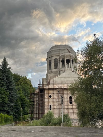 The image shows a large, unfinished concrete structure, likely a religious building, with a prominent dome. The building is surrounded by scaffolding, indicating ongoing construction. Trees and greenery are visible in the foreground and background, suggesting a natural setting. A crane is partially visible in the upper left corner, further emphasizing the construction activity. The sky is filled with dramatic clouds, with patches of sunlight breaking through, indicating either dawn or dusk. The overall mood is one of progress and anticipation, with the imposing structure gradually taking shape against a backdrop of natural beauty. The scene is set in Dilijan, Armenia. There is no visible text in the image.
