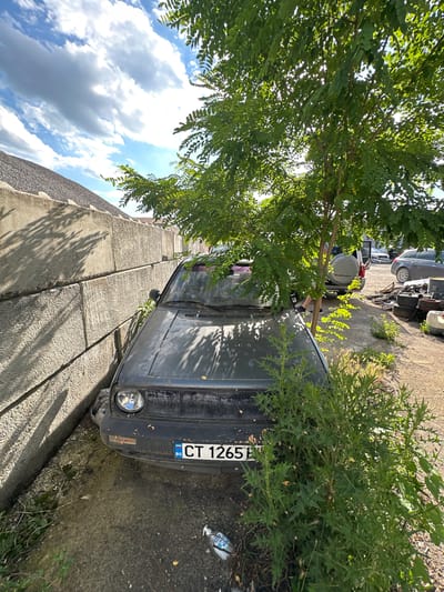 An abandoned, dark-colored Volkswagen Golf Mk2 is parked next to a concrete barrier in an outdoor lot. The car's front end is partially obscured by a leafy tree and dense green bushes. The license plate, BG CT 1265, is visible. In the background, a white SUV with a spare tire on the back, and another car, are visible, along with stacks of scrap materials and tires. The sky is partly cloudy and the scene is illuminated by sunlight, suggesting it is daytime. The location is identified as Kazanlak, Bulgaria.