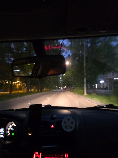 The image is a first-person view from inside a car driving at night on a paved road in Perm, Russia. The dashboard is visible in the foreground, with a smartphone mounted in a holder. To the left, the speedometer and other dashboard lights are illuminated. The road ahead is lit by the car's headlights and streetlights. Trees line the sides of the road, and buildings are visible in the distance. The sky is dark blue, indicating early evening or late twilight. The interior of the car is dark, with only the dashboard lights and reflections from outside providing illumination. The overall atmosphere is one of quiet travel through a city at night.