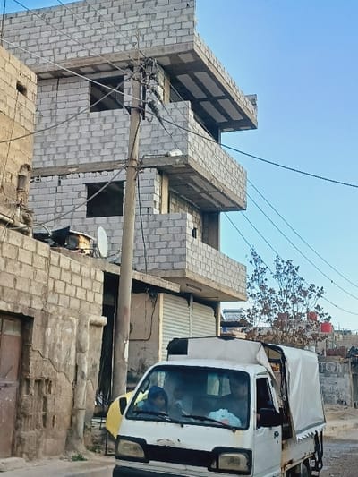 The image depicts an urban street scene in Kafr Sousa Municipality, Syria, under a clear blue sky. A white, visibly aged pickup truck with a white canvas bed cover occupies the lower right and center-front. Two individuals are seated within the truck's cab; the passenger, on the left, wears a blue head covering.
A concrete utility pole stands centrally, laden with numerous black electrical or communication wires and multiple cross-arms. To the left of the pole, an older, multi-story building constructed of light-colored concrete blocks exhibits signs of wear and cracking. A satellite dish is affixed to its roof, and a weathered wooden door is visible at ground level.
To the right of the utility pole, a multi-story building, likely unfinished or under construction, is visible. It is constructed from light-colored concrete blocks, featuring exposed concrete floor slabs serving as balconies or overhangs. Window openings are present but lack frames or glass. The ground level of this structure displays two storefronts with white corrugated metal roll-down shutters.
In the background, behind the truck and further right, a leafless tree and additional residential structures with red rooftop water tanks are discernible. The overall environment suggests a densely built-up area.