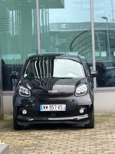 A black Smart EQ electric vehicle is parked facing the camera on a paved surface composed of light-colored interlocking pavers. The car displays a French license plate, "WW-957-VS 67," registered in the Bas-Rhin department, which borders Kehl, Germany. Its front headlights and lower fog lights are illuminated. Directly behind the vehicle stands a modern building with expansive, greenish-tinted reflective glass panels. These panels show reflections of internal structural elements, distant buildings, and faint architectural or industrial structures. A faint reflection of an individual holding a camera or smartphone is visible in the lower right portion of the building's glass facade, positioned above the car's front right quarter panel. The specific street or precise location within Kehl, Germany, is not ascertainable from the visual information. The scene is illuminated by diffused natural light, consistent with an overcast day.